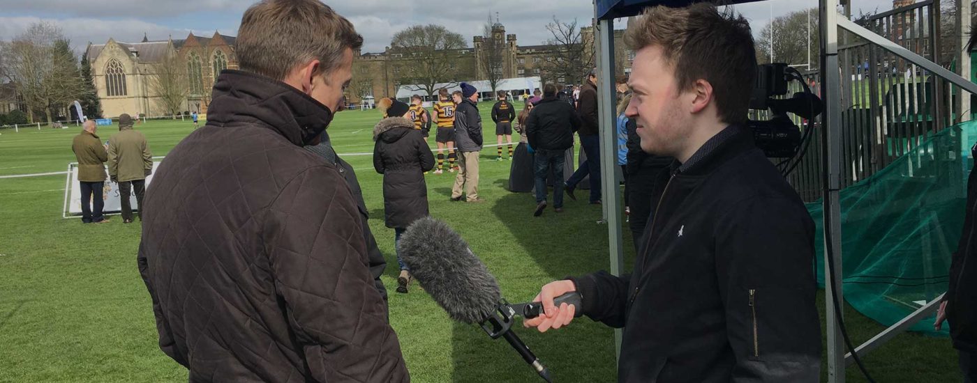Sports journalism student with a deadcat microphone, interviewing a sports person while at a sporting event pitch.