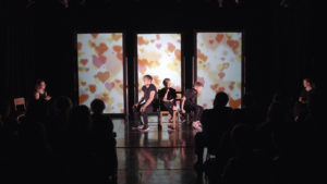 Two actors from the IDEK Theatre Company are sitting on a theatre stage performing. There is a backdrop with large pink and orange hearts. In the foreground audience members can be seen in profile