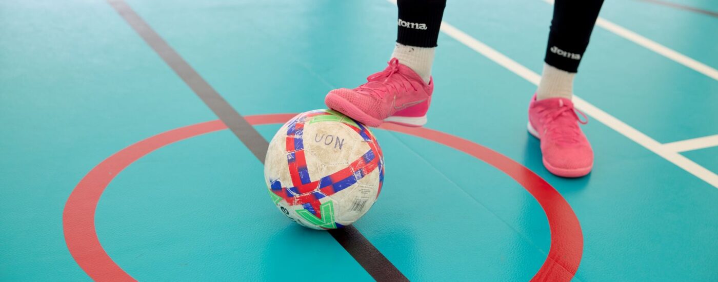 A person wearing pink shoes stands with one foot on a soccer ball inside a red circle on an indoor gym floor.