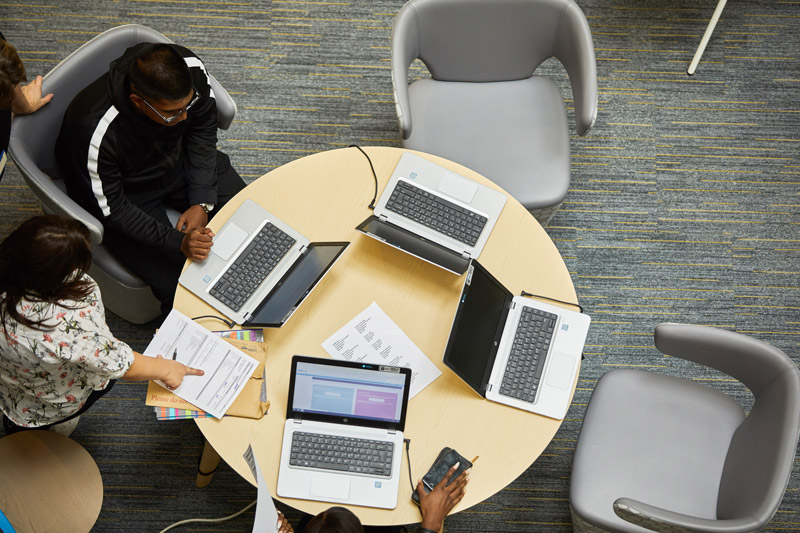 A bird's eye view of two students at a table in the library with a tutor. Their laptops are on the table.