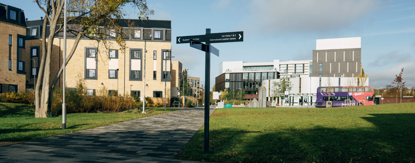 Sign at Waterside Campus on footpath, pointing to student village and leather centre, with the campus behind it