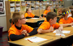 Young children, wearing orange t-shirts sitting at desks with paper in front of them.