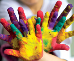Two pairs of children's hands covered in colourful paint, from yellow, purple, green, and pink.