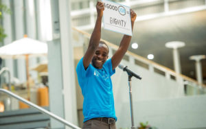 Young person holding a sign that says 'Dignity' in front of microphone at an event. They are smiling