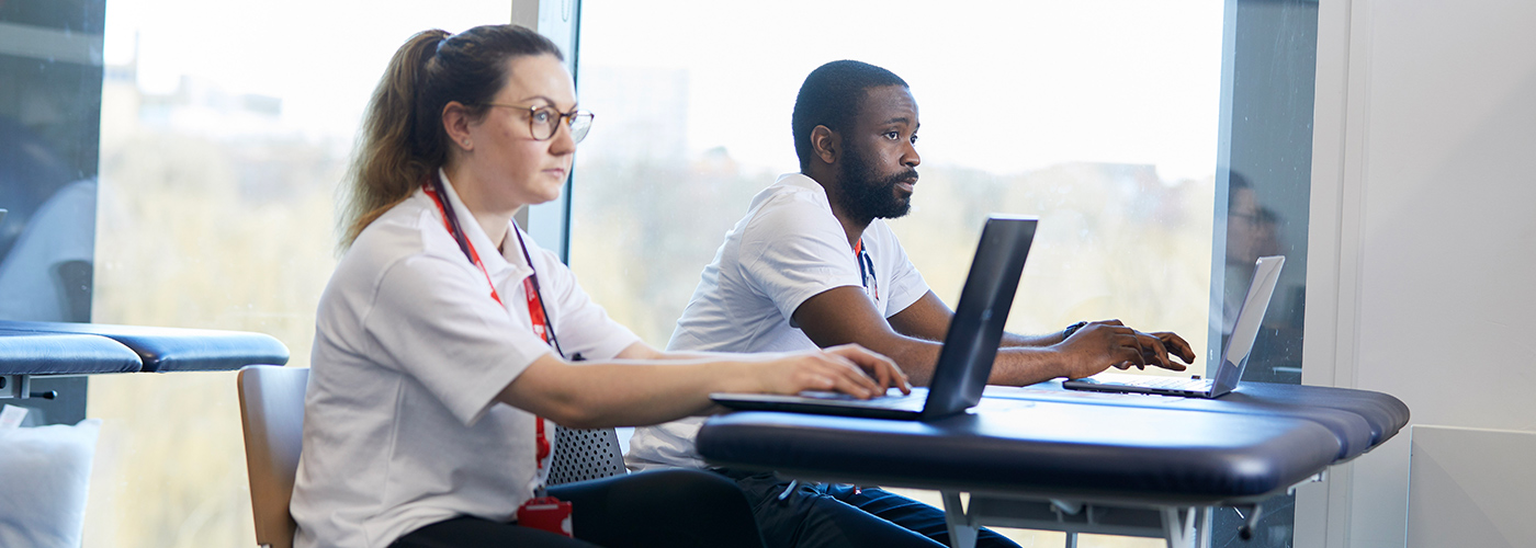 Two physiotherapy students are sitting at their desks on laptops, looking away from the camera towards a tutor.