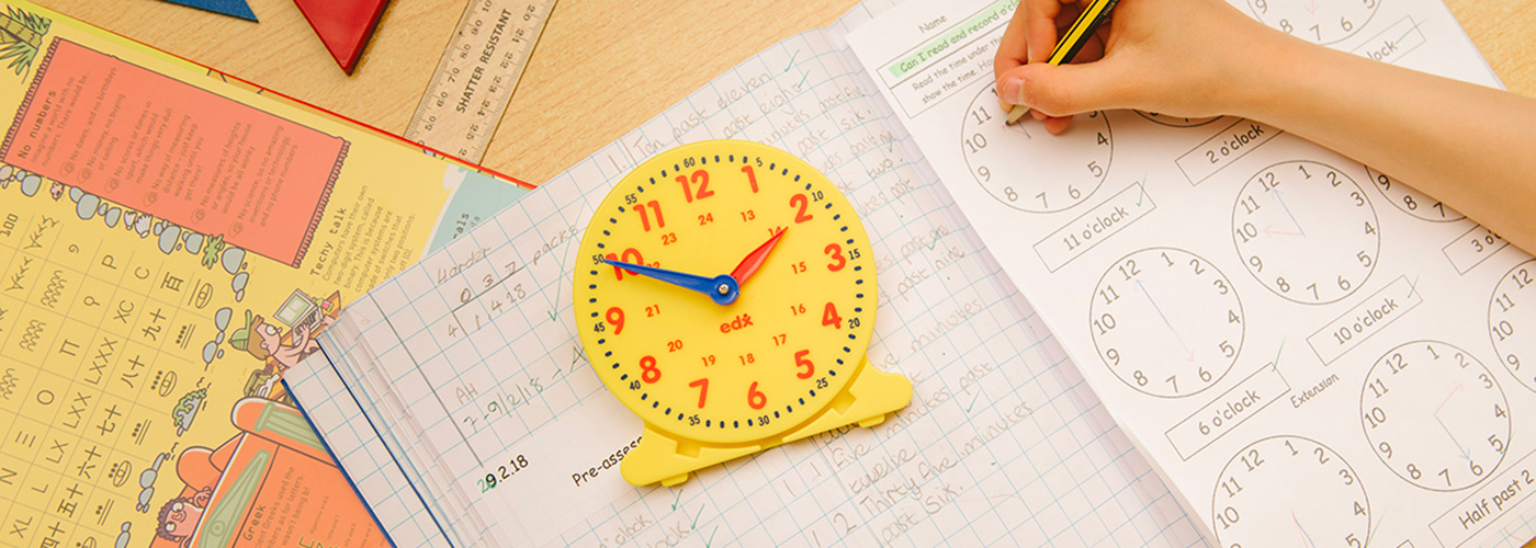 A clock on top of math books, where a child is writing down a clock time in a book.