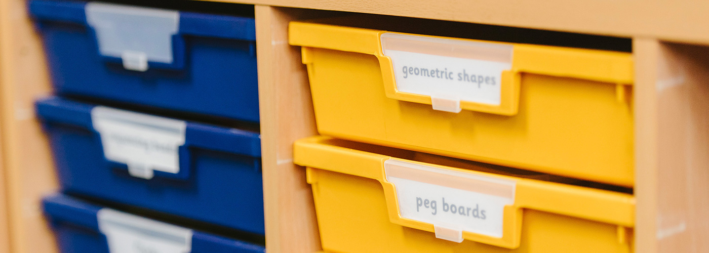 Blue and yellow drawers in a children's classroom. The yellow drawers are labelled for 'geometric shapes' and 'peg boards'.