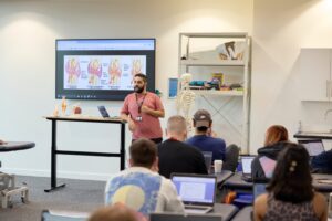 A man gives a presentation on joint anatomy to a classroom of students, with diagrams displayed on a screen and a skeleton model in the background.