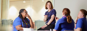 Academic sitting on edge of desk talking to student nurses demonstrating sign language.