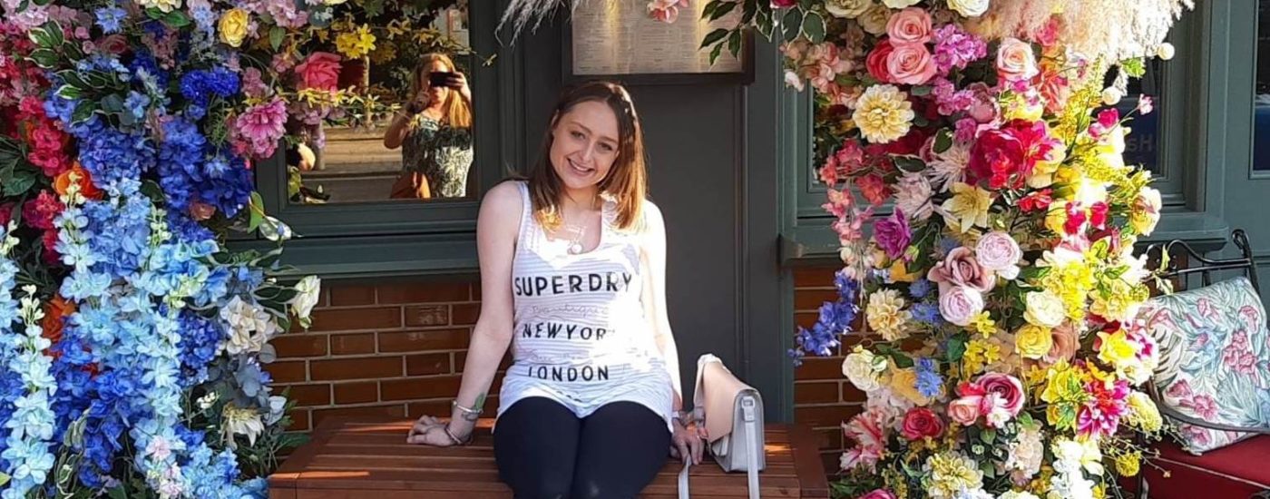 Charlotte Lawton, Mental Health Nursing student, sitting outside of a shop under a colourful flower arch. The flowers on the left are mostly blue, with the ones on the right a mixture of yellow, pink, and green.