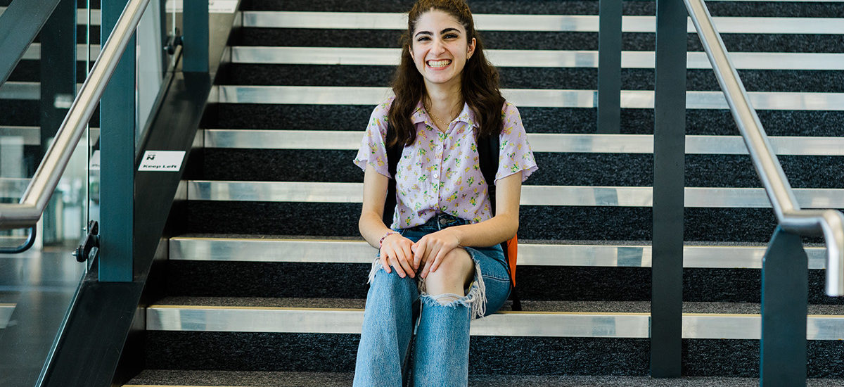 Photo of Health Studies and Applied Social Care student Pel Ahmed. Pel is sitting on the steps inside the Learning Hub, with her hands on her knees and a big smile towards the camera.