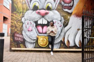 A person in a light hoodie leans against a wall with colorful graffiti featuring a large white cartoon rabbit and holds a smartphone.