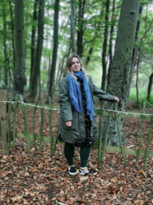 Gemma Robinson-Round, standing in a woods area, next to a wooden fence looking at the camera.