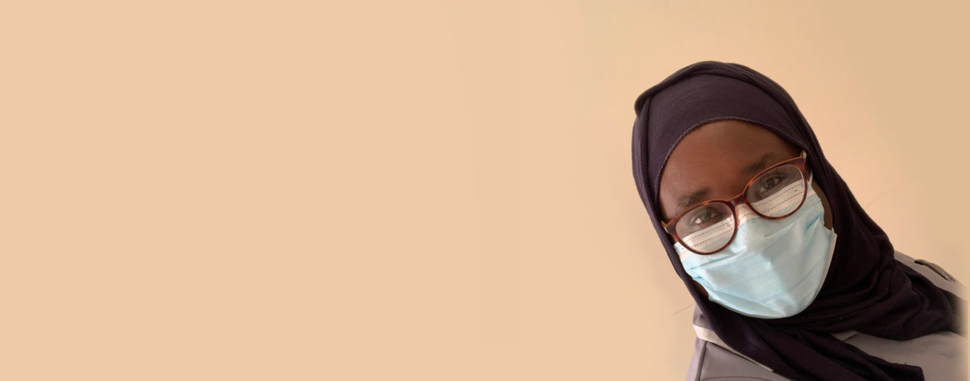 Hauwa Hamza, a Midwifery student, in front of a cream background. Hauwa is wearing a face mask and glass, plus their midwifery uniform.