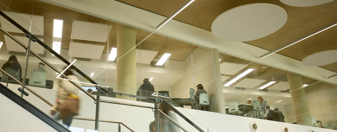 Students seen behind glass windows which show seminar rooms, stairway to first floor of Learning Hub seen in front with students walking up