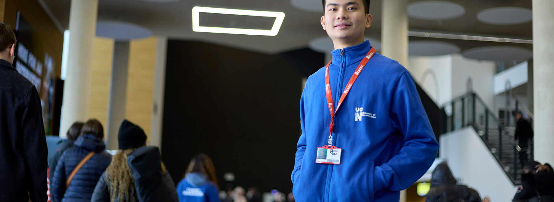 Student ambassador is standing and looking towards the camera, wearing the blue fleece ambassador uniform. Behind them the Learning Hub is full of guests and visitors at an Open Day, all walking away from the camera.