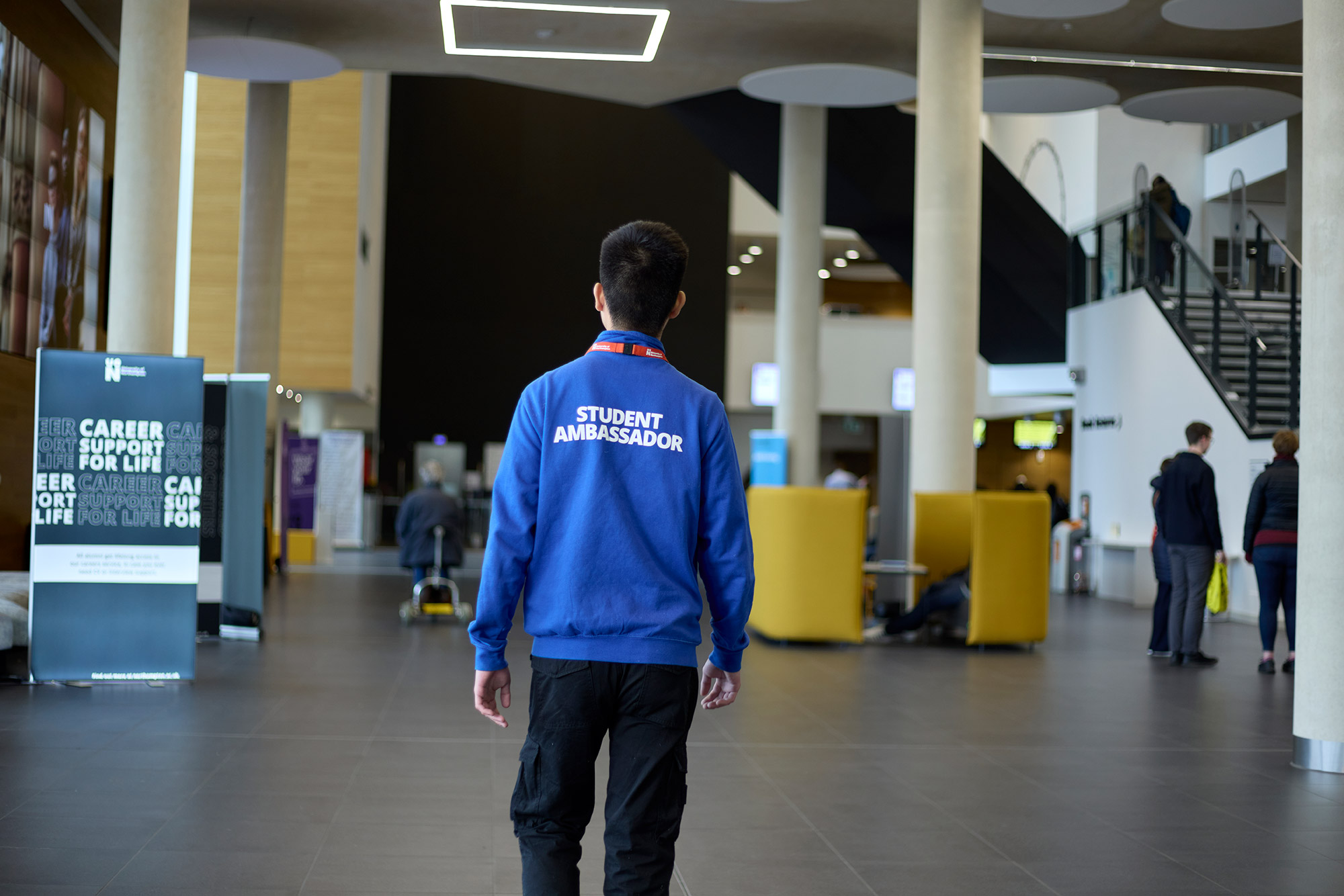 Student Ambassador walking away from the camera, on the ground floor of the Learning Hub. Their blue fleece uniform has 'student ambassador' as text on the back. There are signs around including 'career support for life' around, indicating that it is an open day.