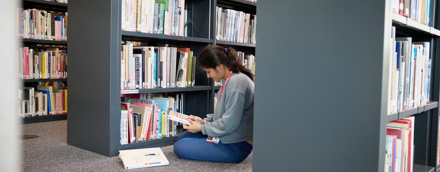 A student is sitting on the floor, wearing jeans and a jumper, holding a book with another next to them, while sitting between three library shelves full of different text books