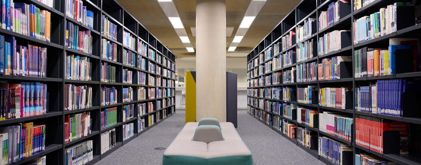Two rows of library shelves extend through the photo, with the camera positioned in the middle showing a shelf on the left and right. In the middle is a bollard, with a long, fabric bench in the middle as a seating area.