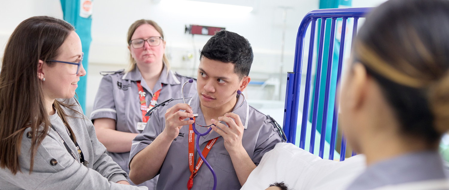 Three student nurses and academic in a practice environment. The student in the middle is wearing a stethoscope, while the academic is demonstrating. Two other students watch. They are in a ward.