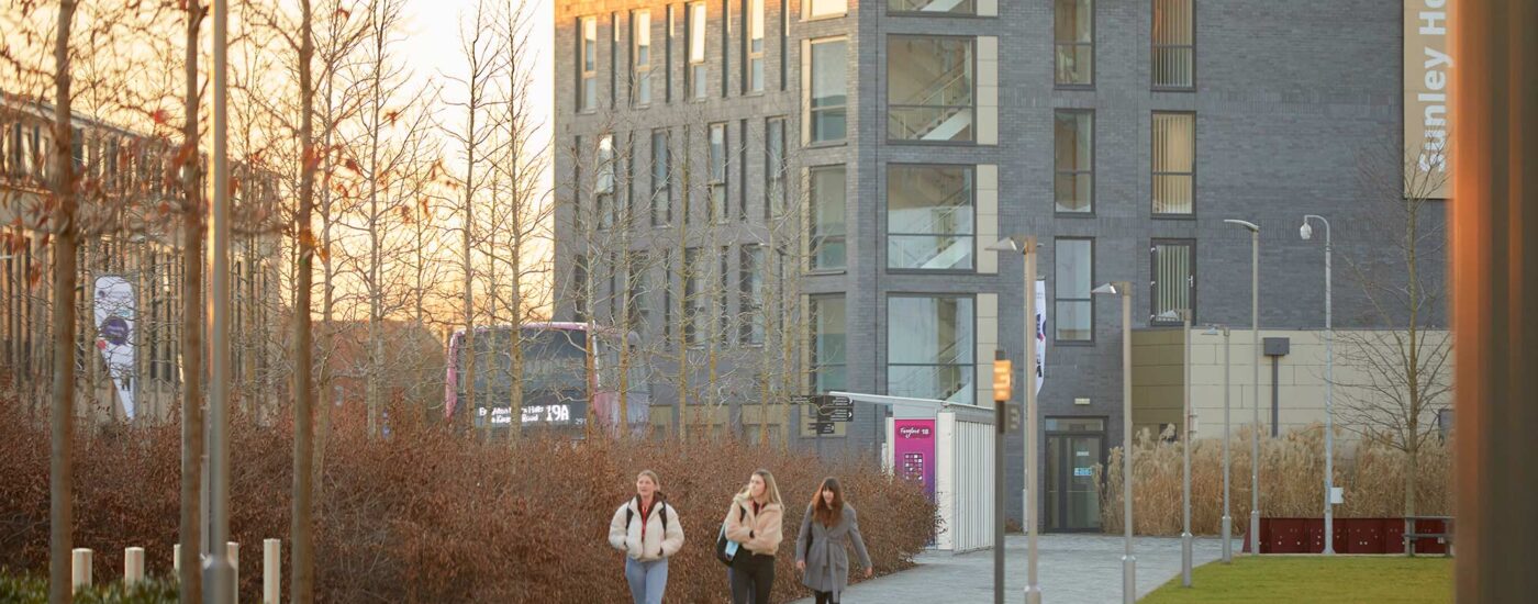 Medium length shot showing three students walking away from bus stop at Waterside campus, in distance.