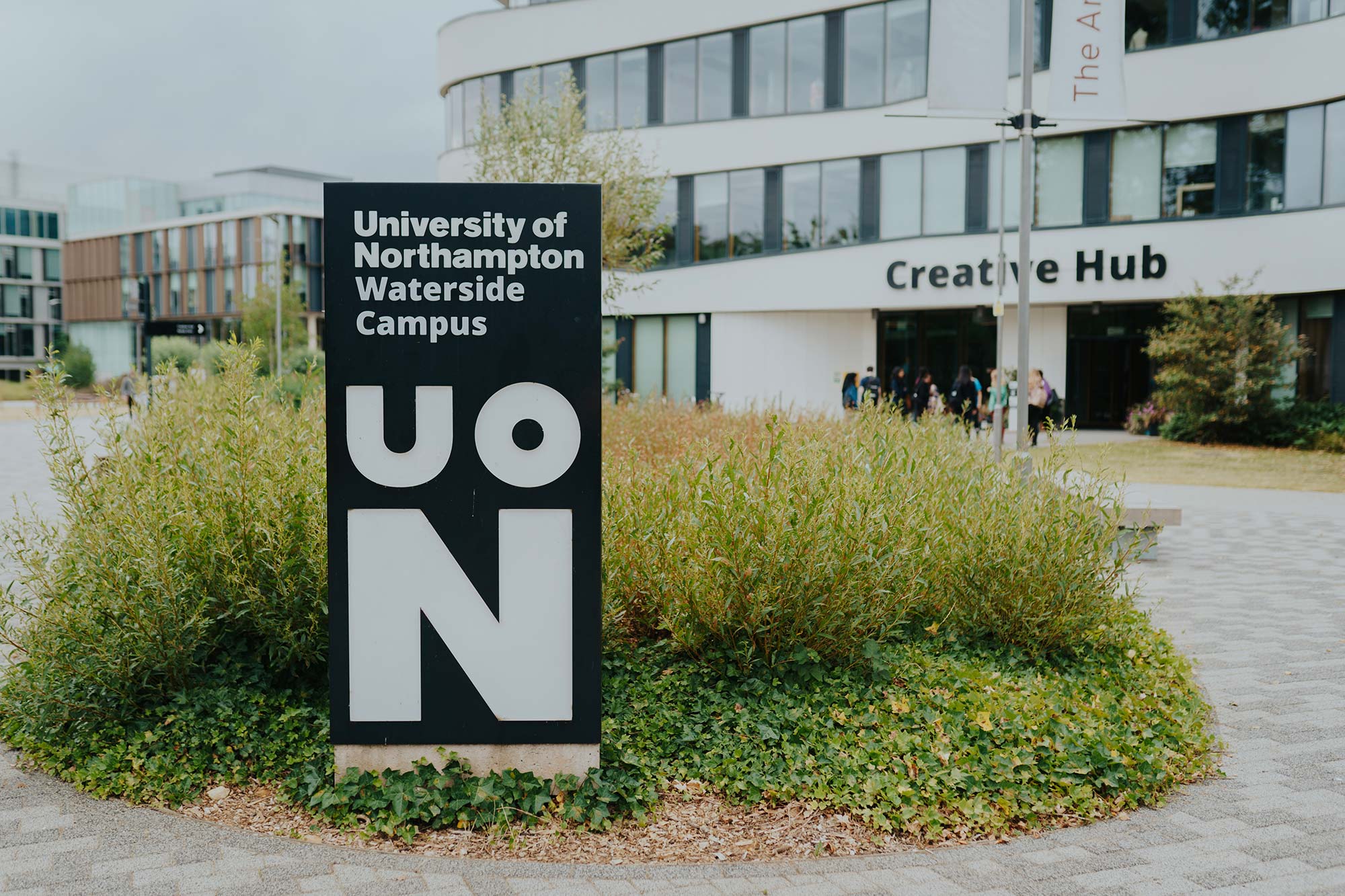 Big sign with 'University of Northampton: Waterside campus' written on it, over UON. The sign is in front of green foliage, which is in front of the Creative Hub building.