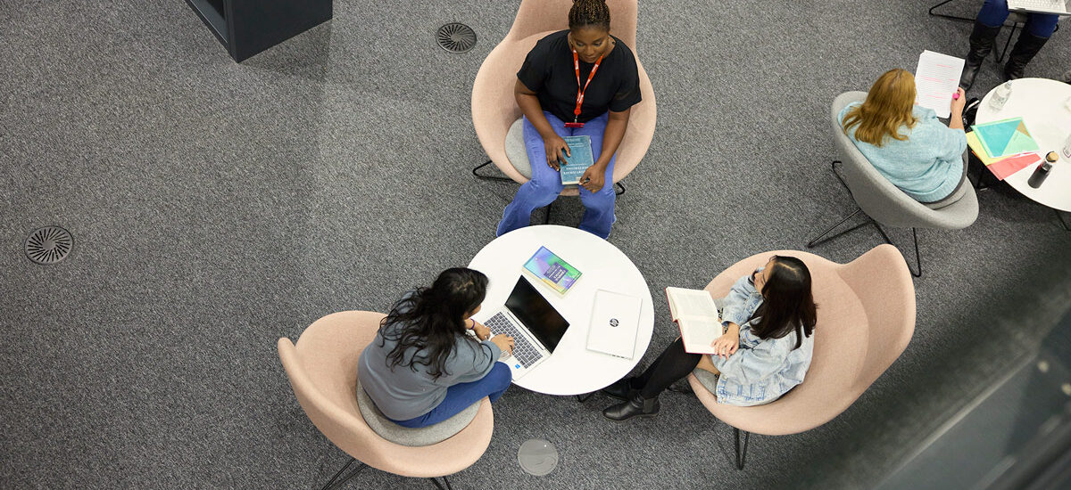A birds eye view of three students sitting in big pink chairs, with a circle table in front of them with their laptops on. One student has a book on their lap.