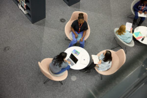 A birds eye view of three students sitting in big pink chairs, with a circle table in front of them with their laptops on. One student has a book on their lap.