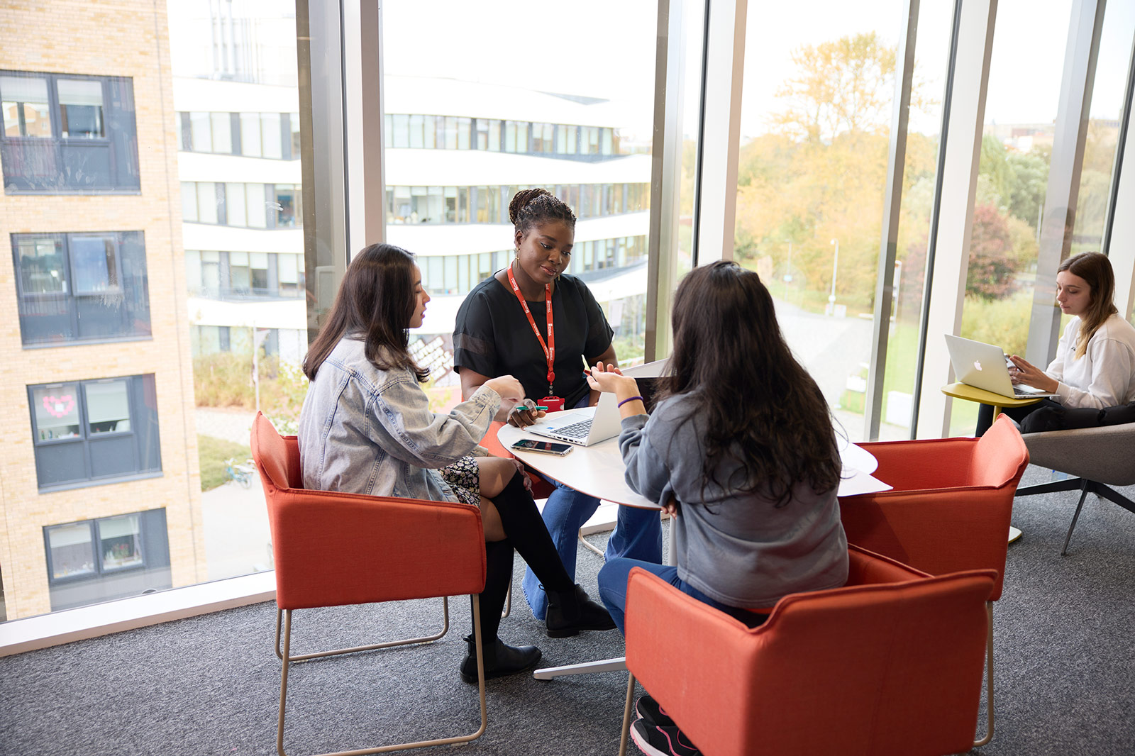 Three students sitting in small orange chairs at a circle table, where one student has a laptop, but the other two are talking. They are in front of a ceiling to floor window, where the Creative Hub and some Accommodation Halls at the University of Northampton can be seen.