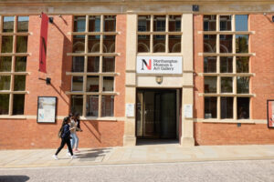 Entrance of the Northampton Museum and Art Gallery, where three students are walking past. The building is brick and the entrance has glass doors. It is a sunny day.