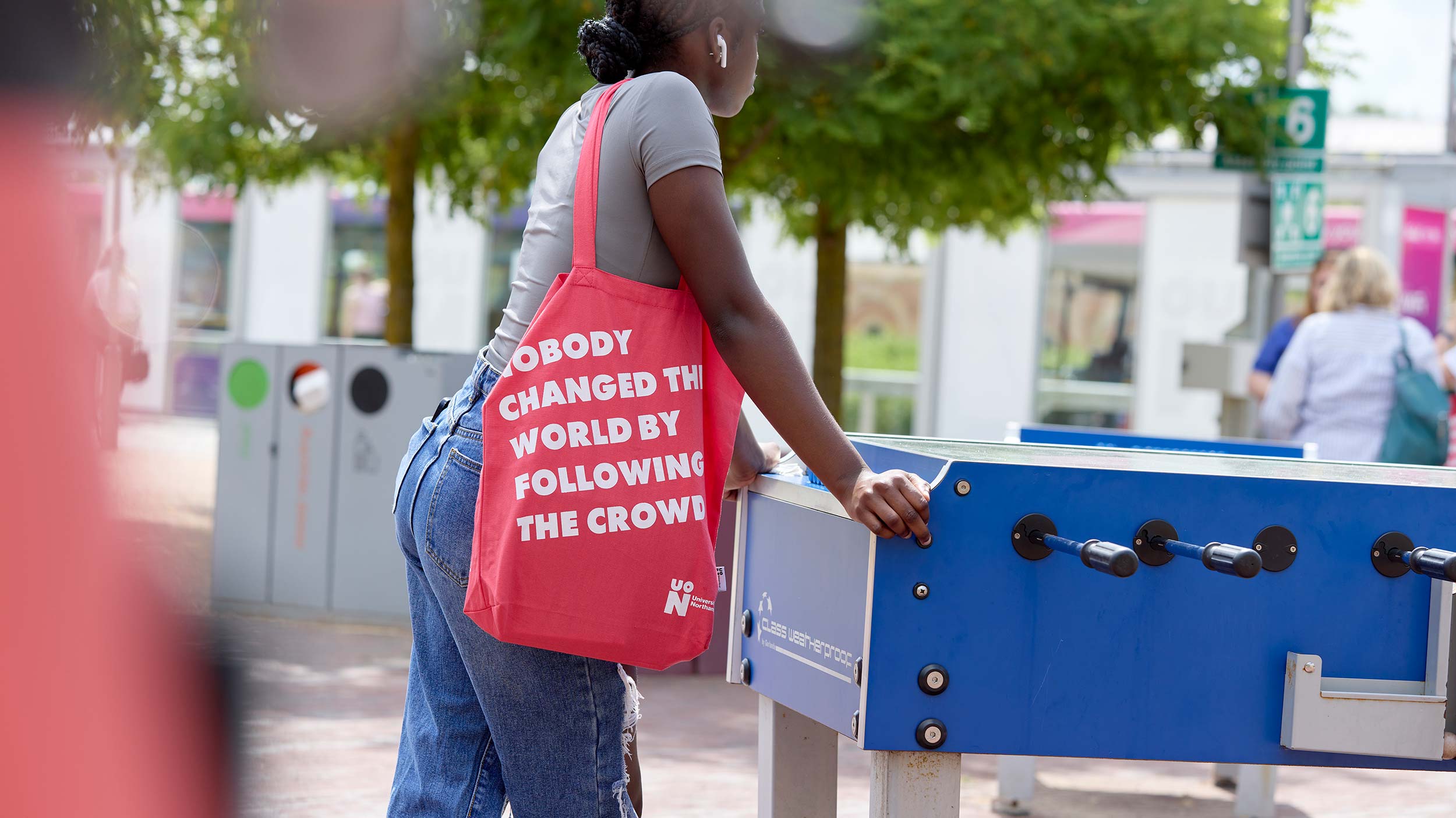 Student standing at a table football station outside. Their face is not in view, but their hands are on the table and they have a red tote bag from a University of Northampton event on their arms which reads 'nobody changed the world by following the crowd'.
