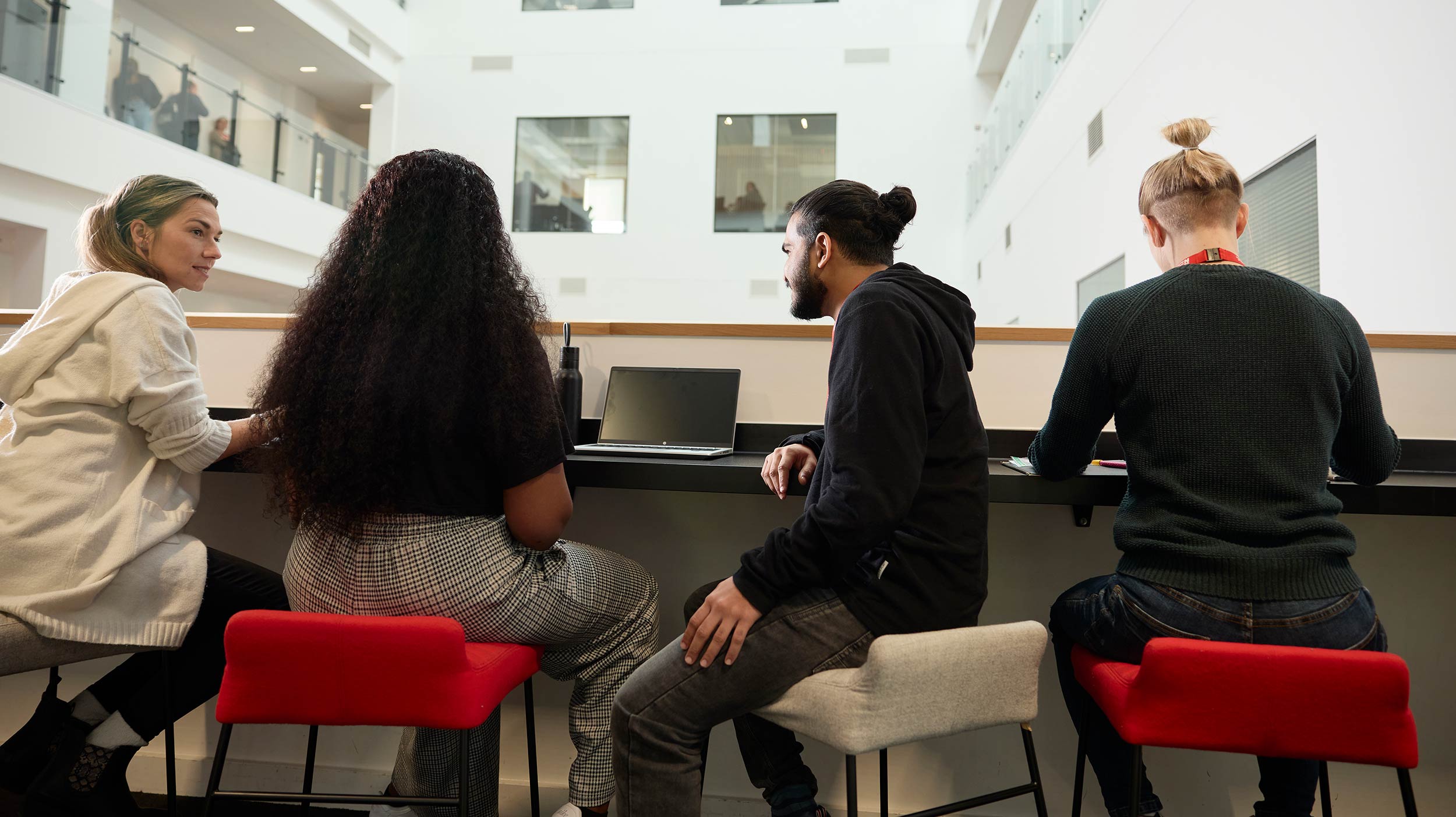 Four students sit on stools in the informal working space in the Senate building on the University of Northampton campus. They have laptops on the ledge in front of them and look out over the Senate atrium with glass walled teaching spaces in the background