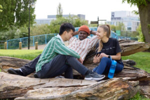 Three students sitting on benches made of wood in Becket's Park, with University of Northampton buildings behind. One student is showing the other two their phone, and they are smiling.