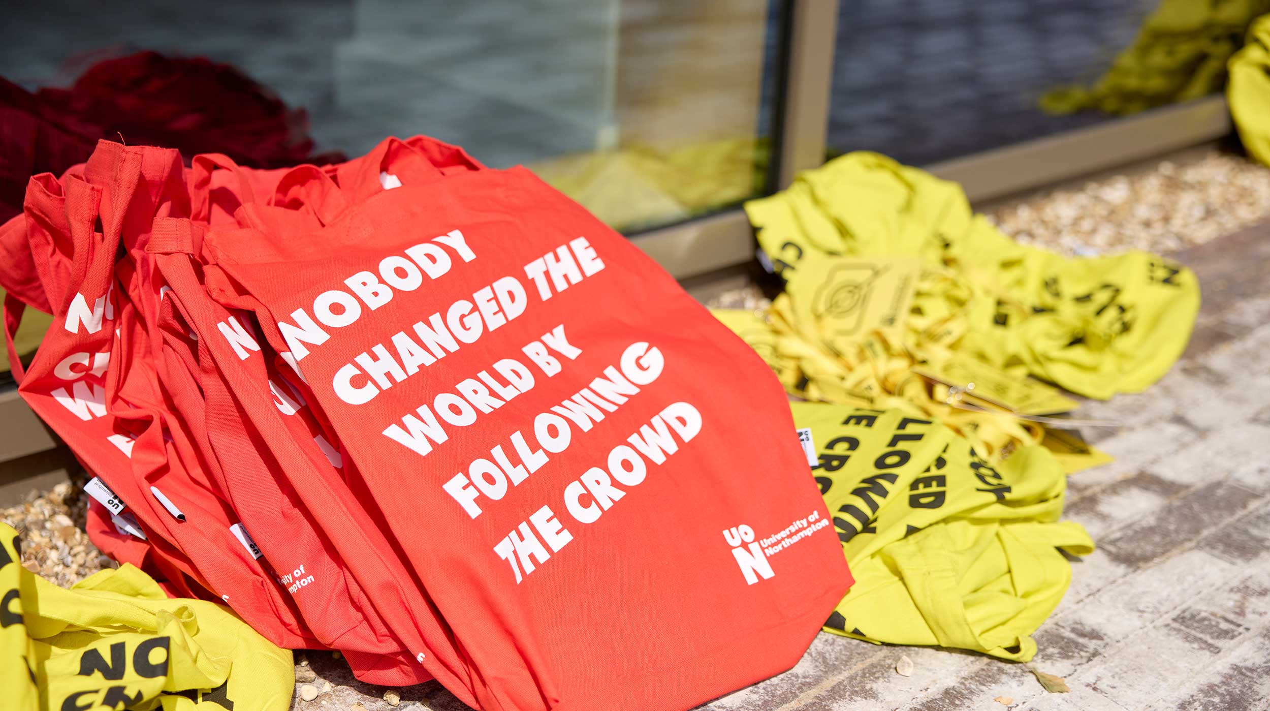 Red cotton tote bags are stacked ready to be handed out to visitors at an open day at the University of Northampton. The bags are printed with the words 'Nobody changed the world by following the crowd'