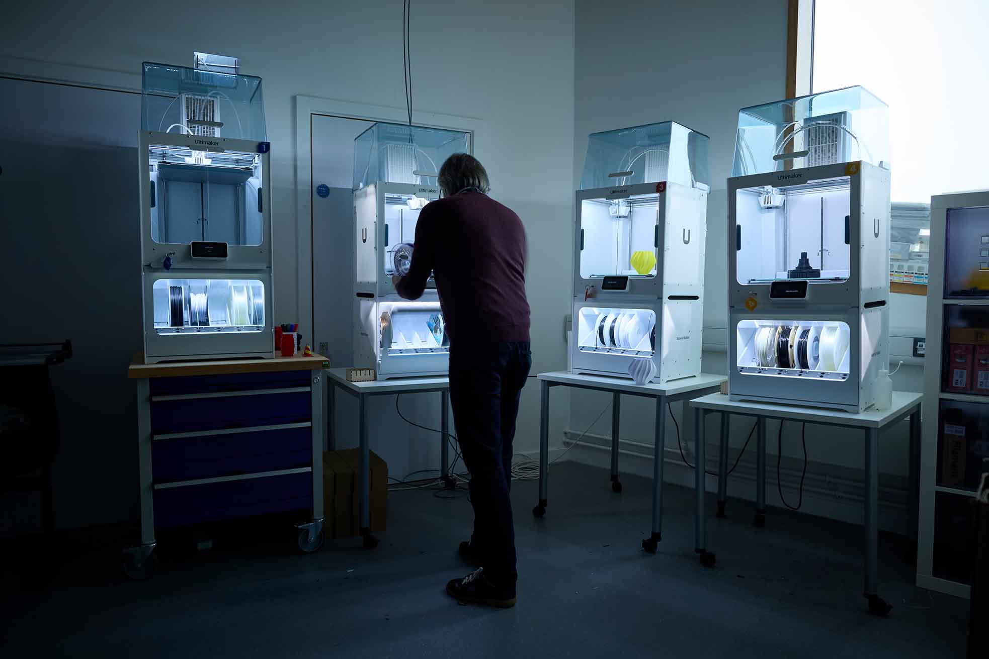The photo shows the 3D printing workshop at the University of Northampton. A student operates one of four print machines with brightly lit screens on each