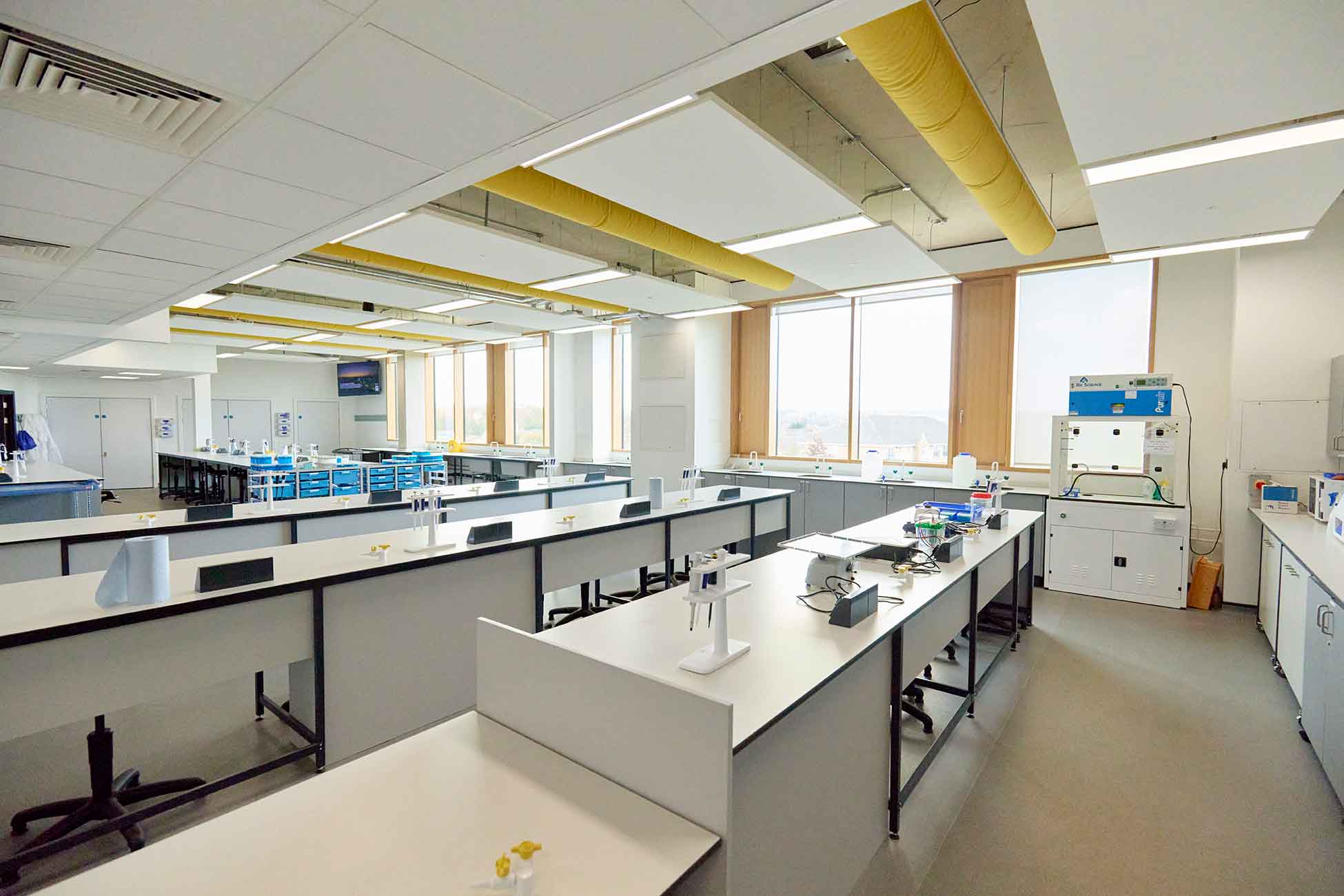 A photograph of the Bioscience Laboratories at the University of Northampton. There are three modern white workbenches with equipment laid out for experiments and floor-to-ceiling windows in the background