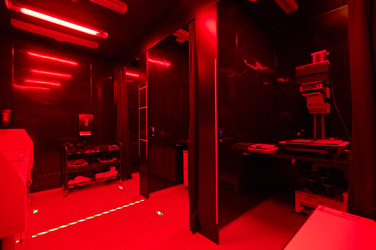 Internal view of the Dark Room, which is a black room with red lights which are reflected off the floors, walls and devices around. There are three rooms with a curtain that can be closed around it, in order to print the photos.
