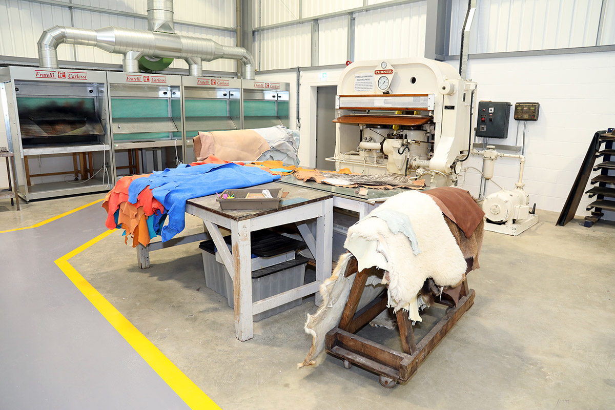 Internal view of the Finishing Room in the Institute Creative Leather Technologies. There is a workbench , with different colour pieces of leather material over them from blue, orange, red, and white. There are machines around the warehouse style room.