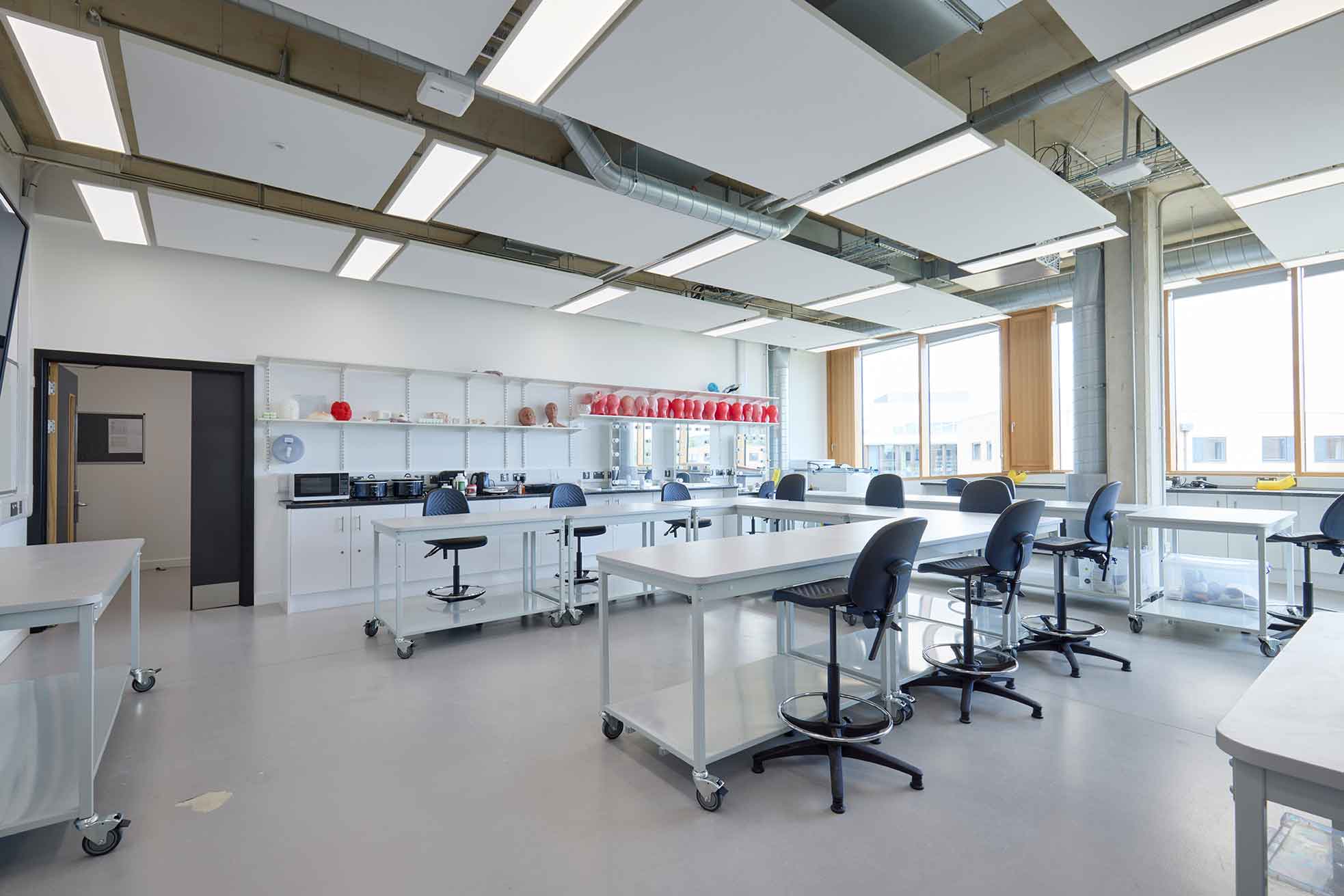 The photo shows the Hair, Makeup and Prosthetics studio in the Creative Hub at the University of Northampton. In the background are floor-to-ceiling windows and in the foreground are swivel stools and workbenches. There are several pieces of specialist palette mixing equipment and a row of cosmetology mannequins arranged along the back wall in the photograph