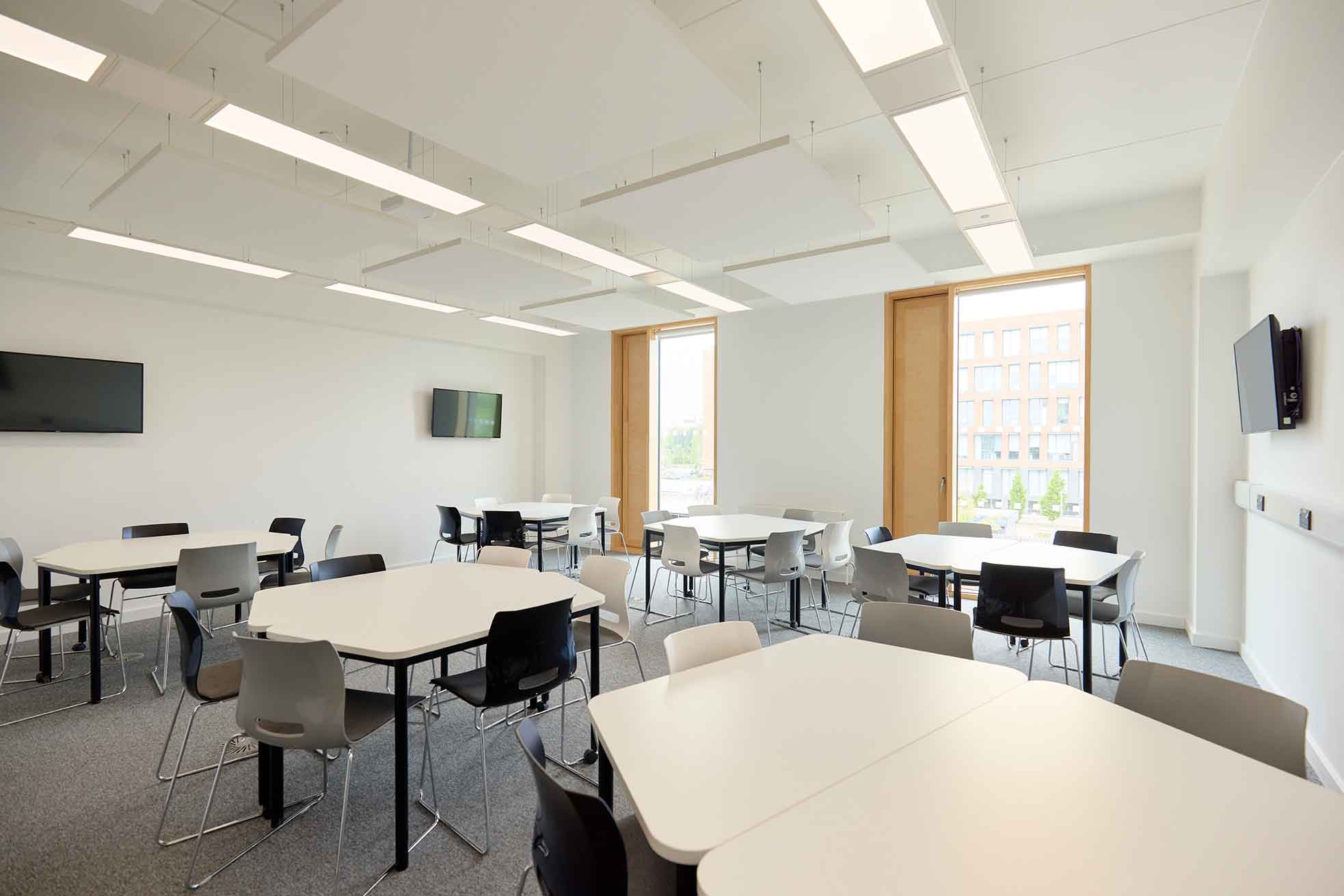 The picture shows a classroom in the Learning Hub with large white tables and a mixture of grey, black and white chairs. There are three digital monitors on the walls and floor-to-ceiling windows
