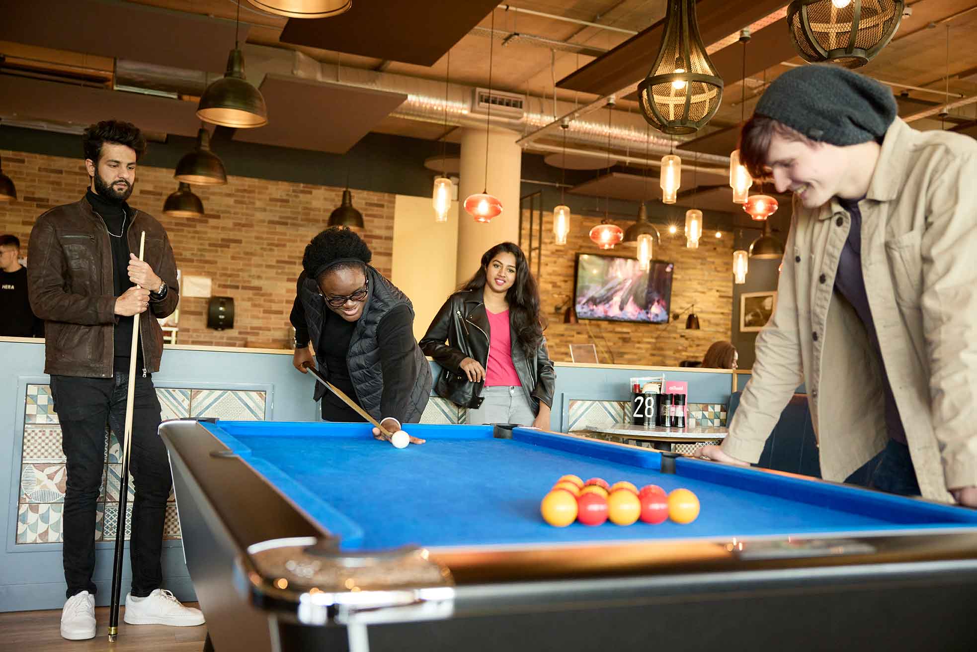 Four students play a game of pool together in the Waterside Bar and Restaurant on campus at the University of Northampton. One student holding a cue takes a shot at the white cue ball while the other three watch