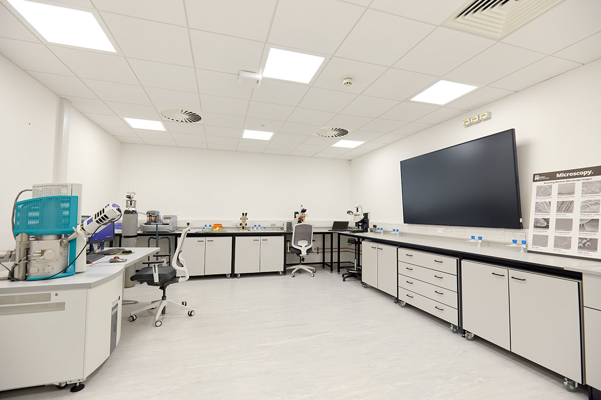 Internal view of the Microscopy Laboratory, which is a white room with cupboards in an L shape. There is a big screen on the right, with microscopes on the benches on the other sides of the wall.