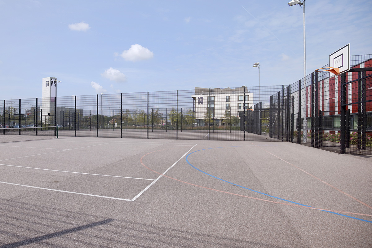 External view of the multi-use games area, which is an outdoor pitch which is encaged with a blank fence. The floor is light grey tarmac, with white, red, and blue lines on it to indicate the different pitches. There is a basketball hoop at one end. In the distance the Energy Centre and Senate building can be seen.