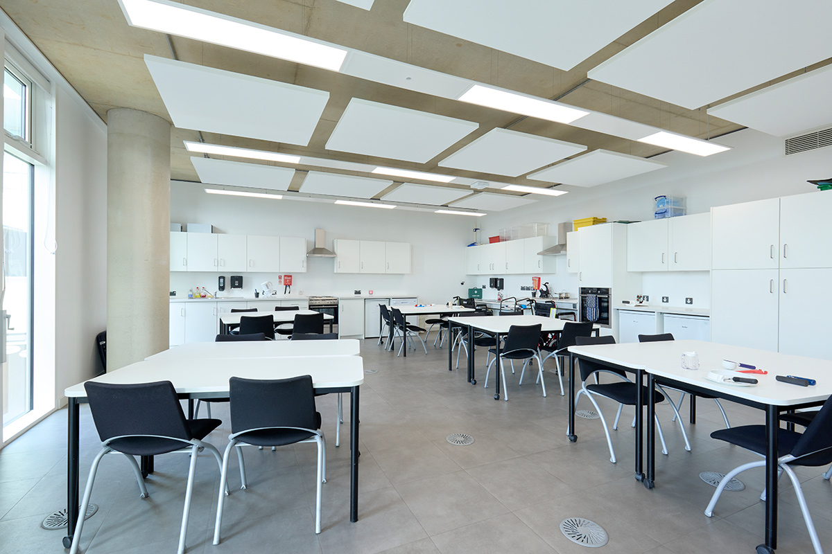 Internal view of the Occupational Therapy rooms, which have tables and chairs that seat four at a table, while the room is surrounded by cupboards and kitchen whitegoods (including oven, fridges, freezes, and a hob)