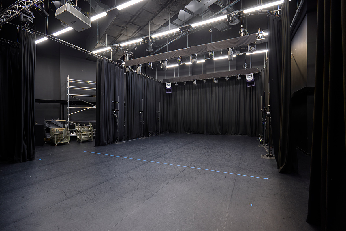 Internal view of the Performance Area, which is a black room with big lights in the ceiling and black curtains to separate into rooms and provide acting capacity. The floor is black, with a blue line across it, to provide a staging area.