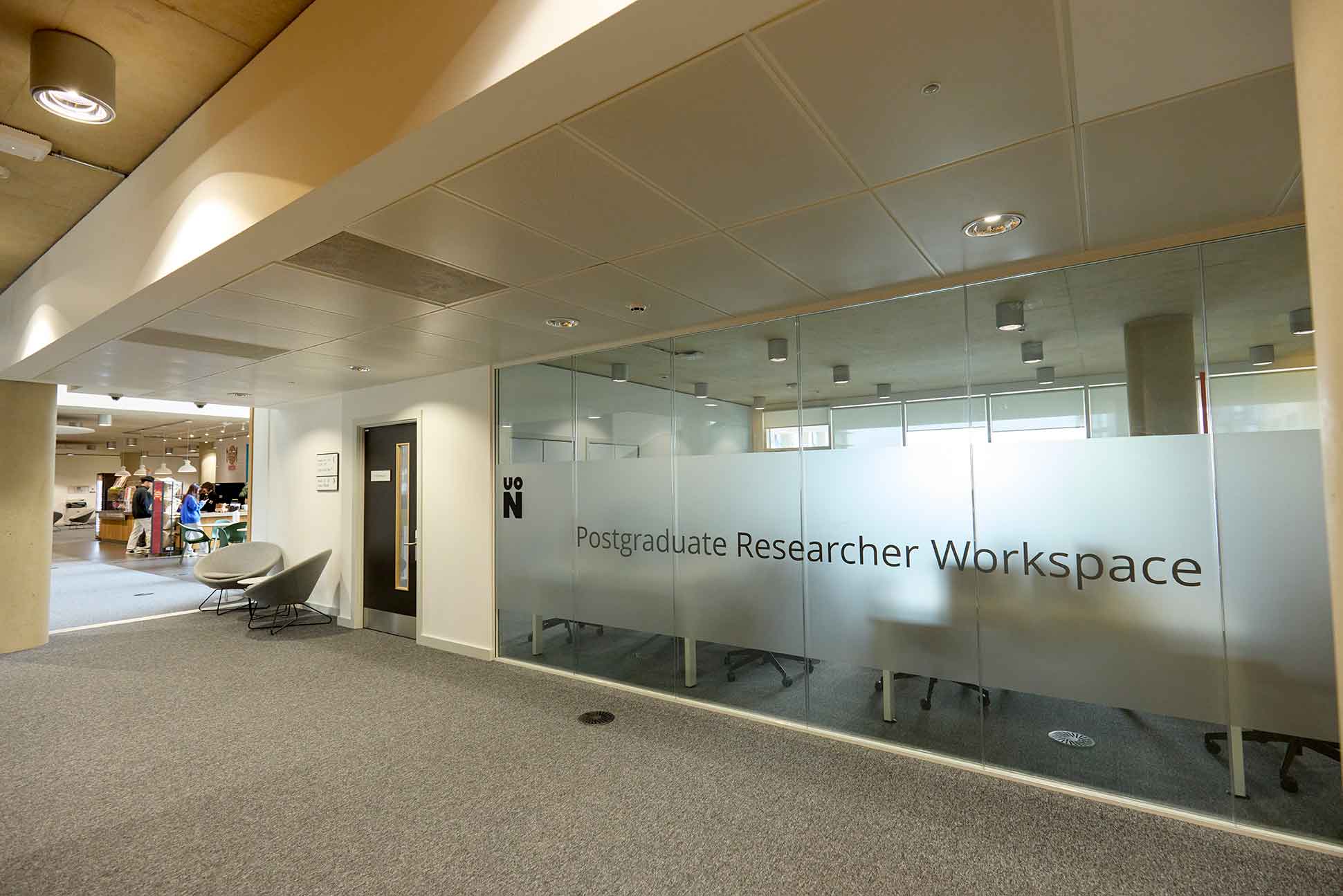 The picture shows the glass wall exterior of the Postgraduate Research Workspace. Through the frosted glass walls there are the outlines of desks and chairs and a window beyond