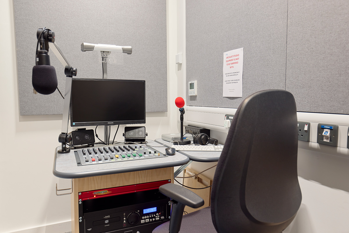 Internal view of the Radio Studio, which shows a corner of the room with a small desk that rests a screen, as well as buttons and switches for recording with a microphone hanging down in front of the desk chair. Under the desk is recording technology, with other buttons on it.