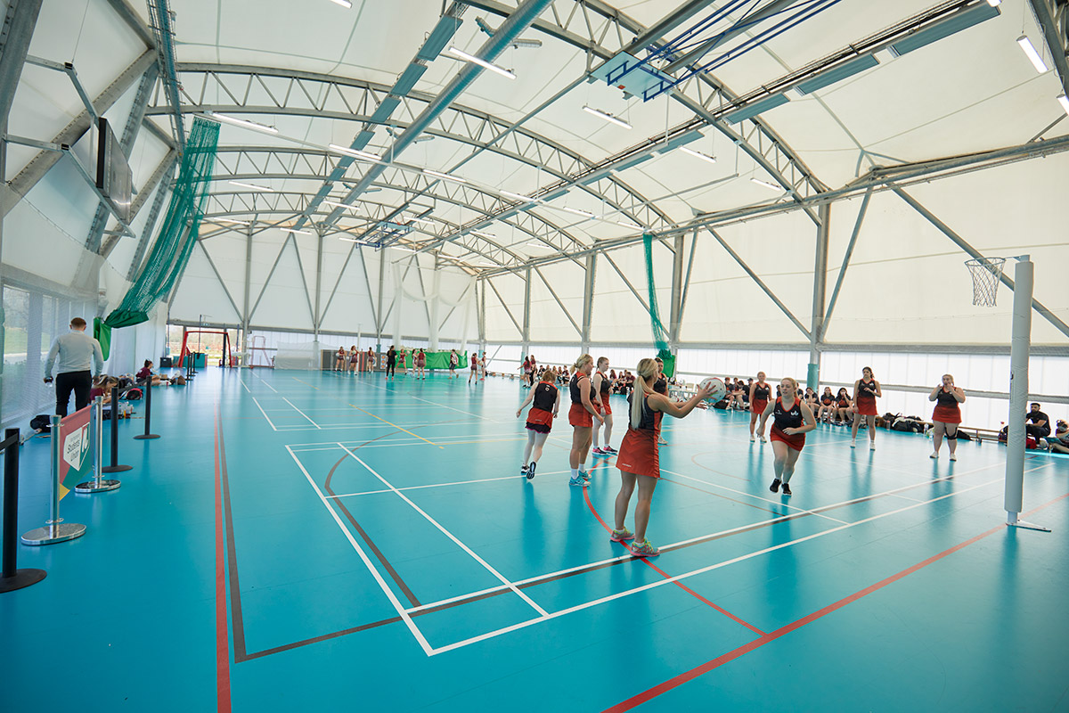 Internal view of the Sports Dome where a netball game is being played. Inside the dome is blue flooring with white, black, and red lines to indicate the different pitches. There are two netball nets on either side and the photo is taken from one end, where activity is happening during the game. One team is holding the ball, aiming for the goal.