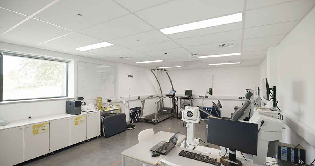 The photo shows the sports laboratory - the room is white with grey flooring and a window to the left of the room. There are monitors and laptops in the foreground and in the background there are sports monitoring machines