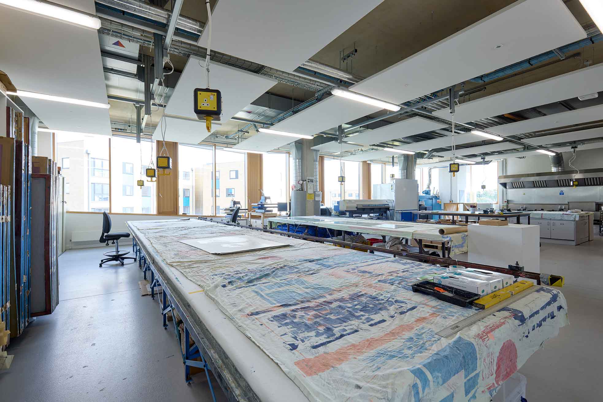 The photograph shows the textile printing workshop at the University of Northampton. In the foreground textile prints are laid out on workbenches in the background are floor-to-ceiling windows and printmaking equipment